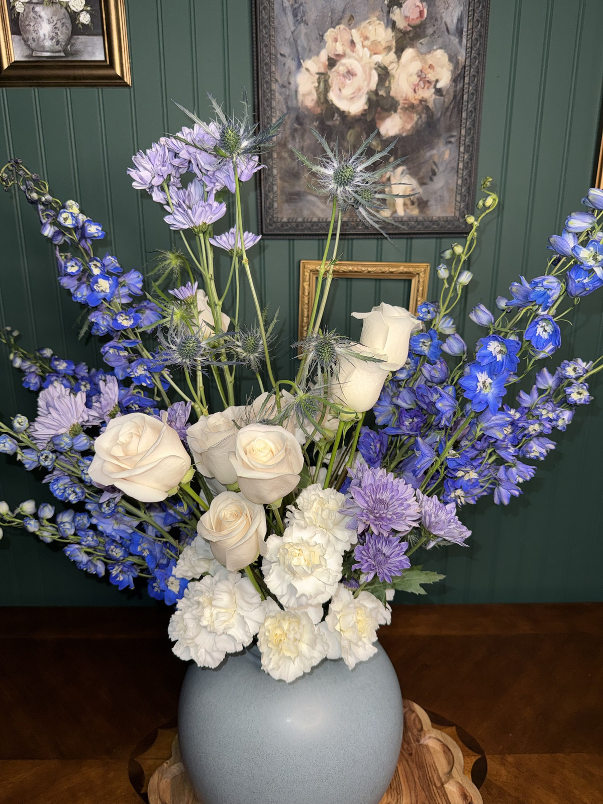 White roses and carnations with blue delphinium and thistle in a round ceramic vase