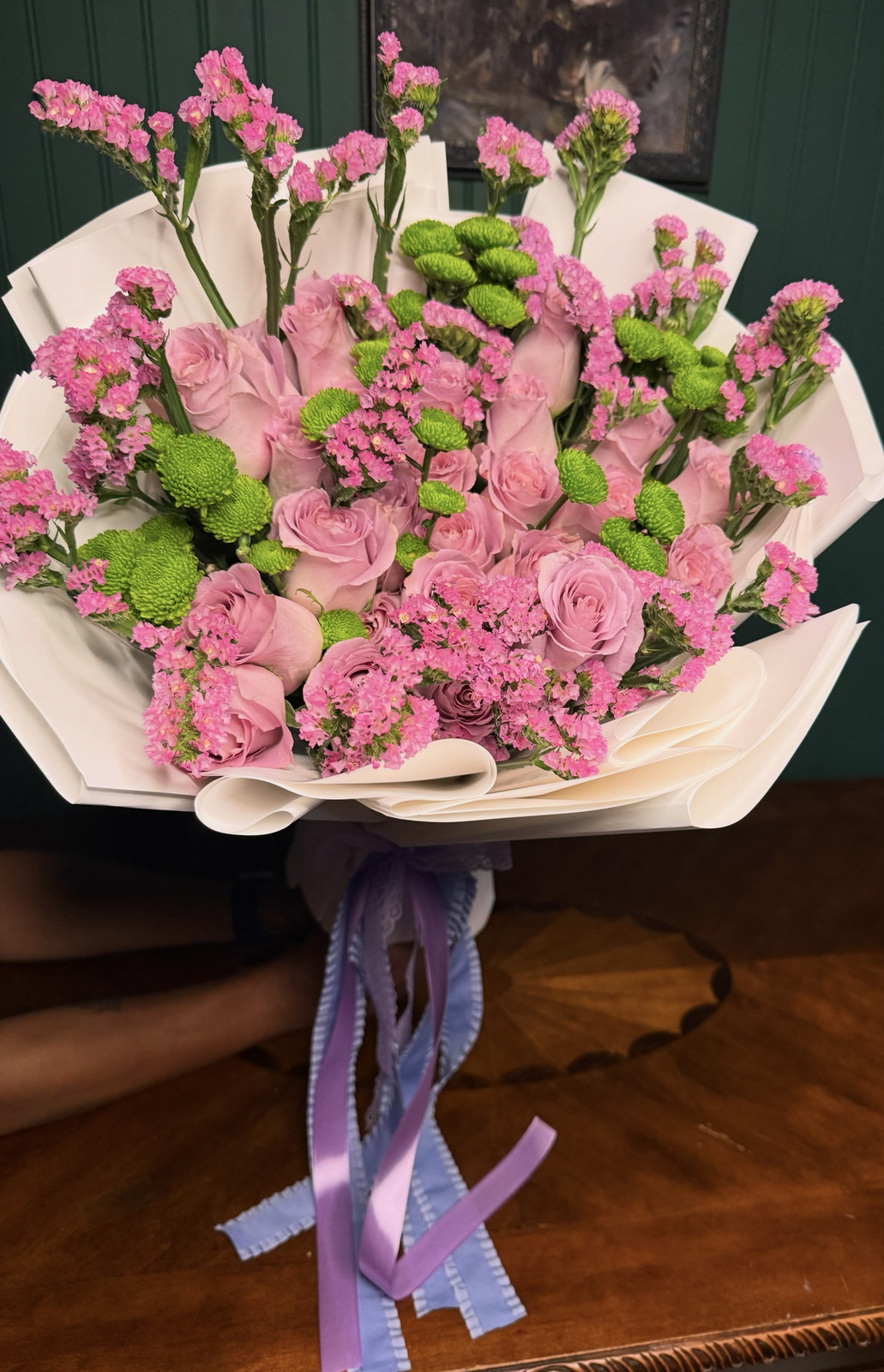 Pink roses, pink statice and green button mums wrapped in white with lilac ribbons