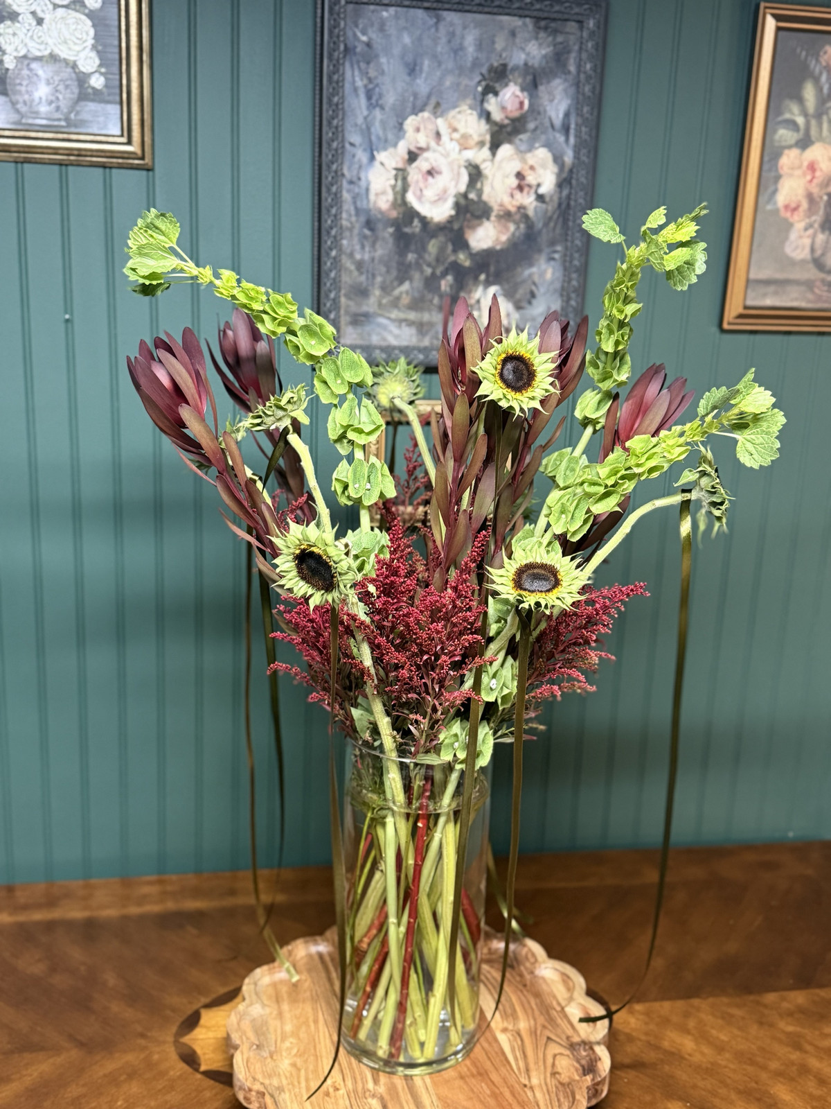 Bells of Ireland, green sunflowers, burgundy leucadendron and red solidago in a tall glass vase