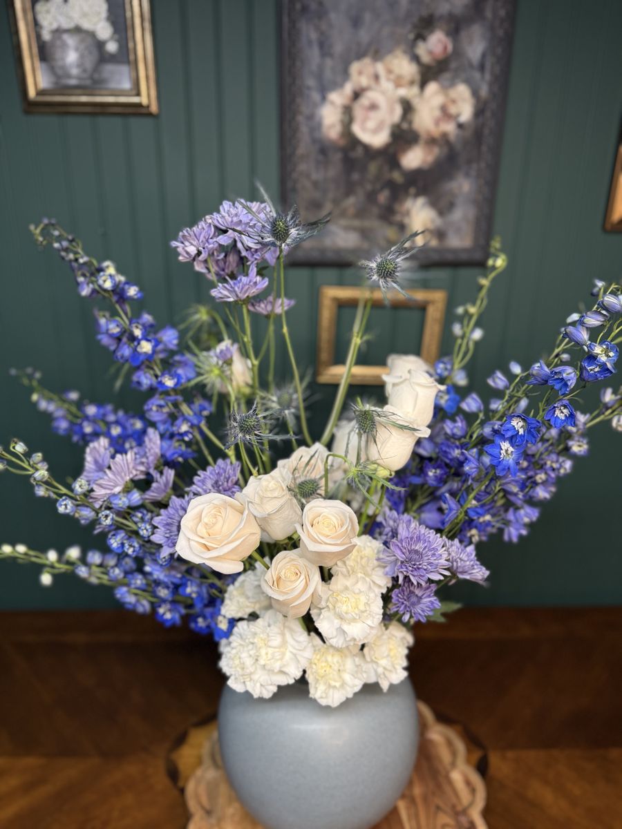 Blue delphinium, white roses, carnations and lavender mums in a round blue vase